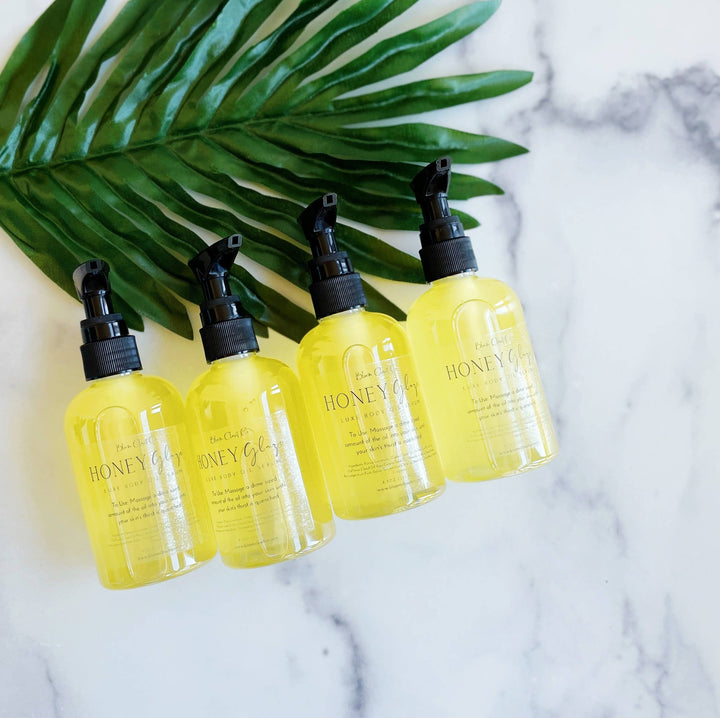 Four bottles of 4oz Honey Glaze Body Oil Serum displayed alongside a palm leaf.